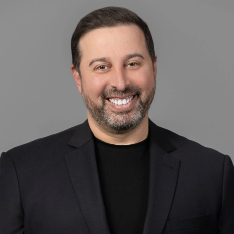 Smiling man in a black shirt and blazer posing against a gray background, a professional headshot. Laid-back, approachable demeanor.