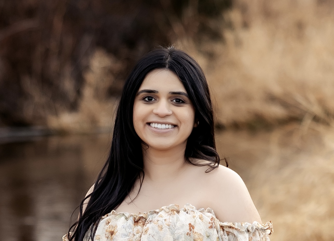 Smiling woman with long dark hair in a floral off-the-shoulder top, outdoors by a blurred brownish background lake.
